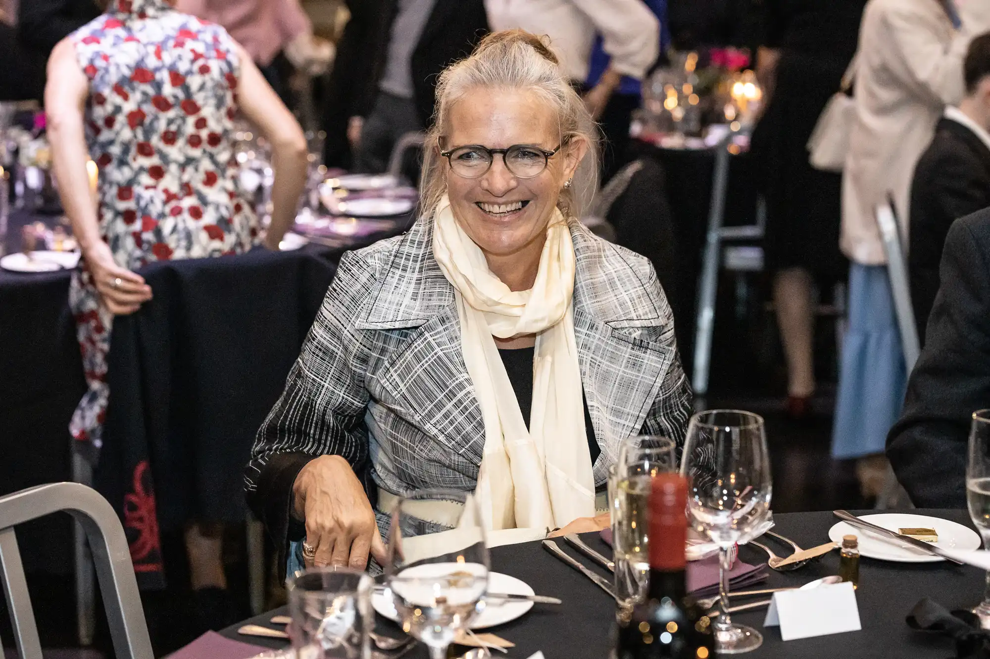 A smiling woman with glasses and a scarf sits at a dinner event. She is surrounded by tables with plates, glasses, and wine bottles. People in the background are engaged in conversation.