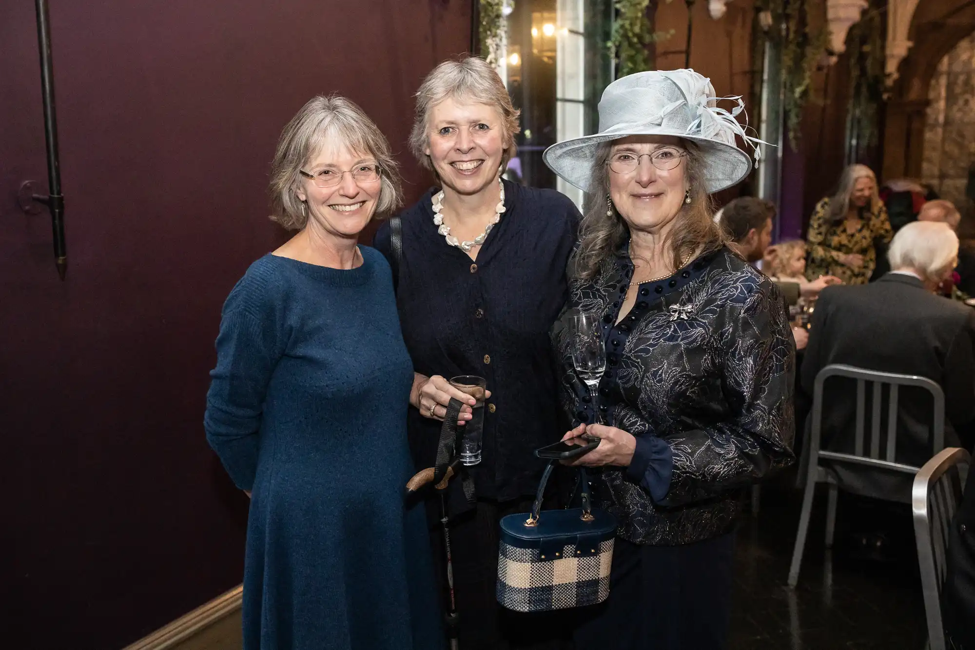 Three women are standing closely together and smiling at an indoor event. They are dressed in semi-formal attire, with one wearing a hat and holding a wine glass. The background features tables, chairs, and guests.