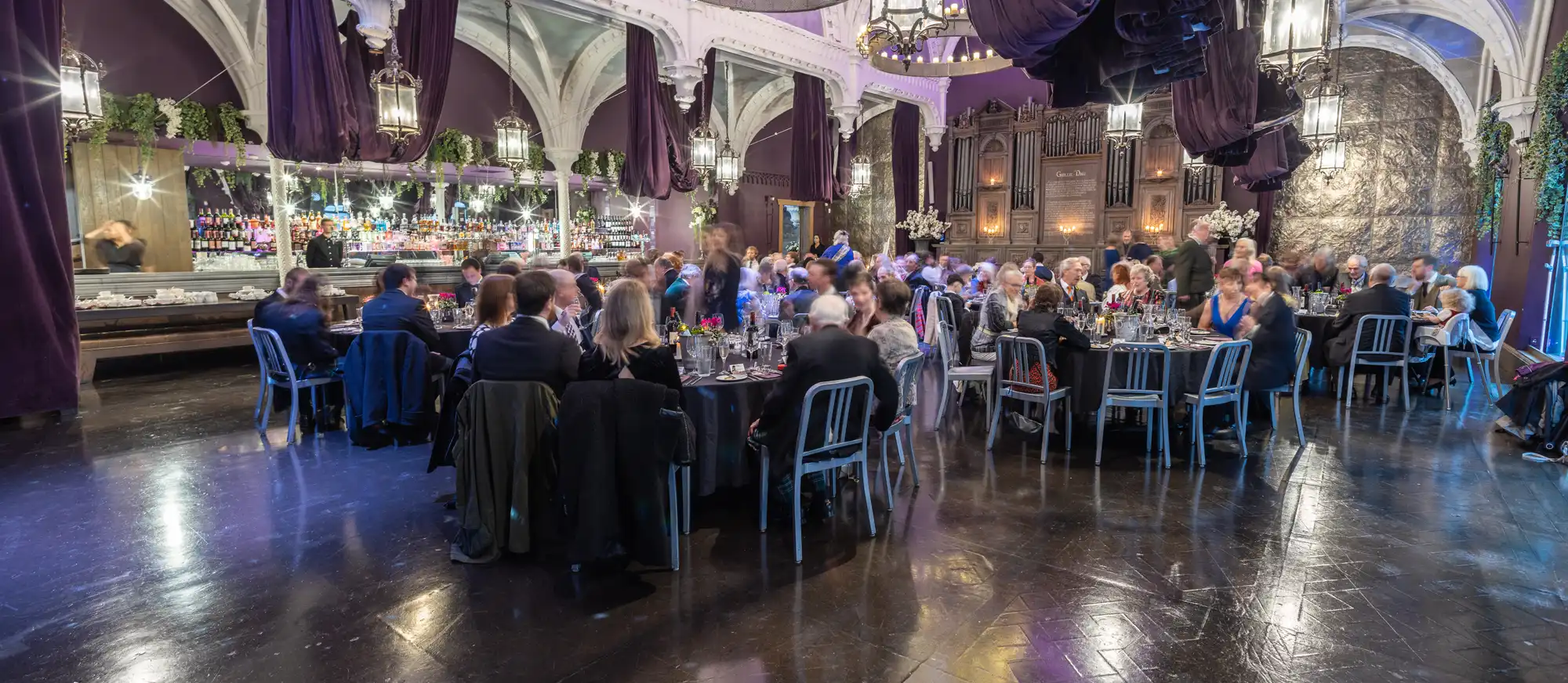 A formal gathering with many people seated at round tables in an elegant hall with high ceilings, chandeliers, and ornate decor.