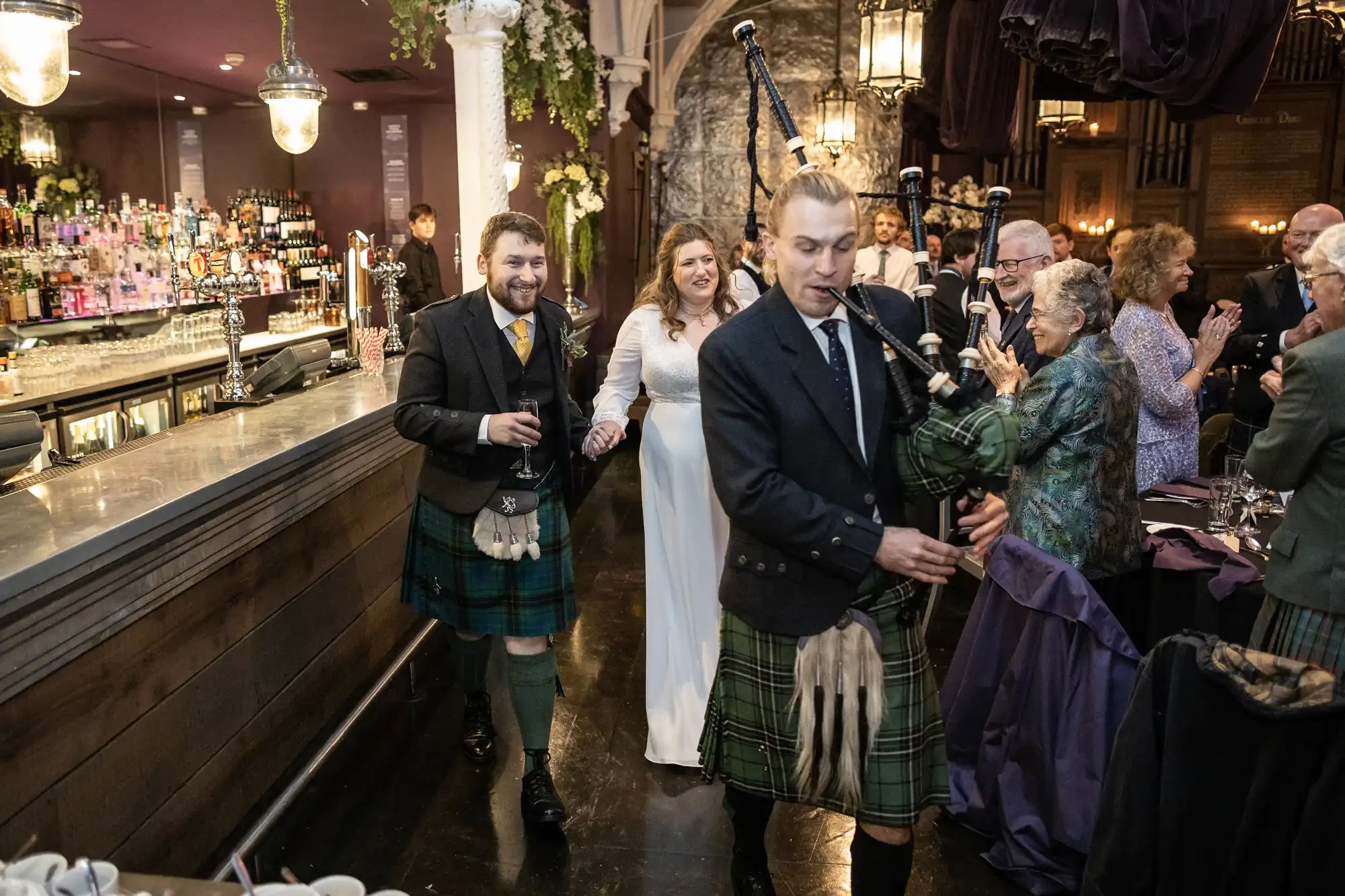 A bagpiper leads a wedding procession in a festive indoor setting, followed by the bride and groom wearing traditional Scottish attire. Guests stand and celebrate near a long bar counter.