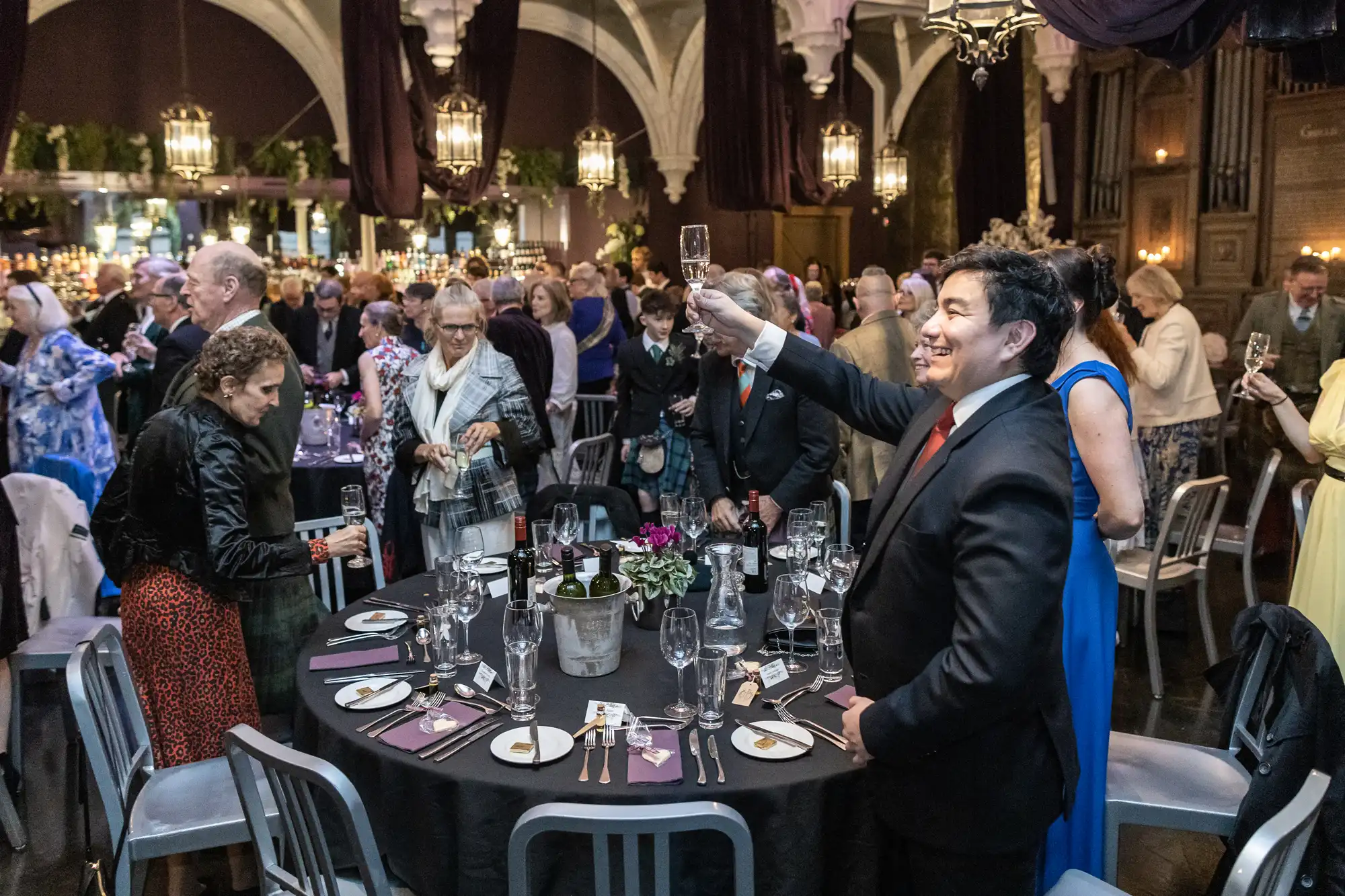 A man raises a glass in a toast at a formal event with attendees gathered around tables set for dining. The venue features high ceilings and hanging lanterns.