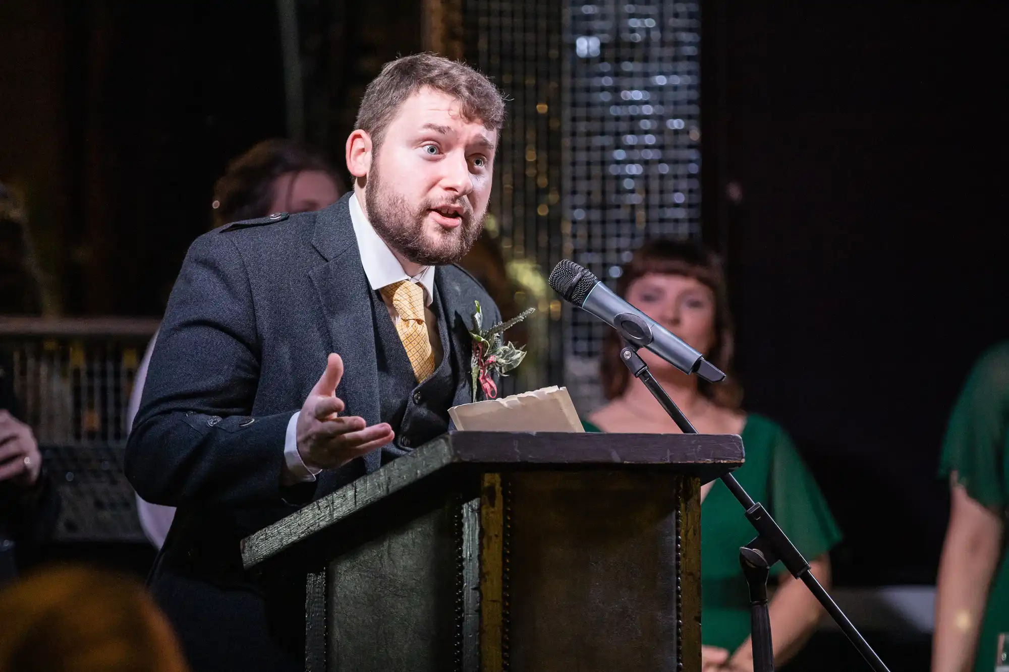 A man in a suit speaks passionately into a microphone at a podium, while people in green attire stand in the background.