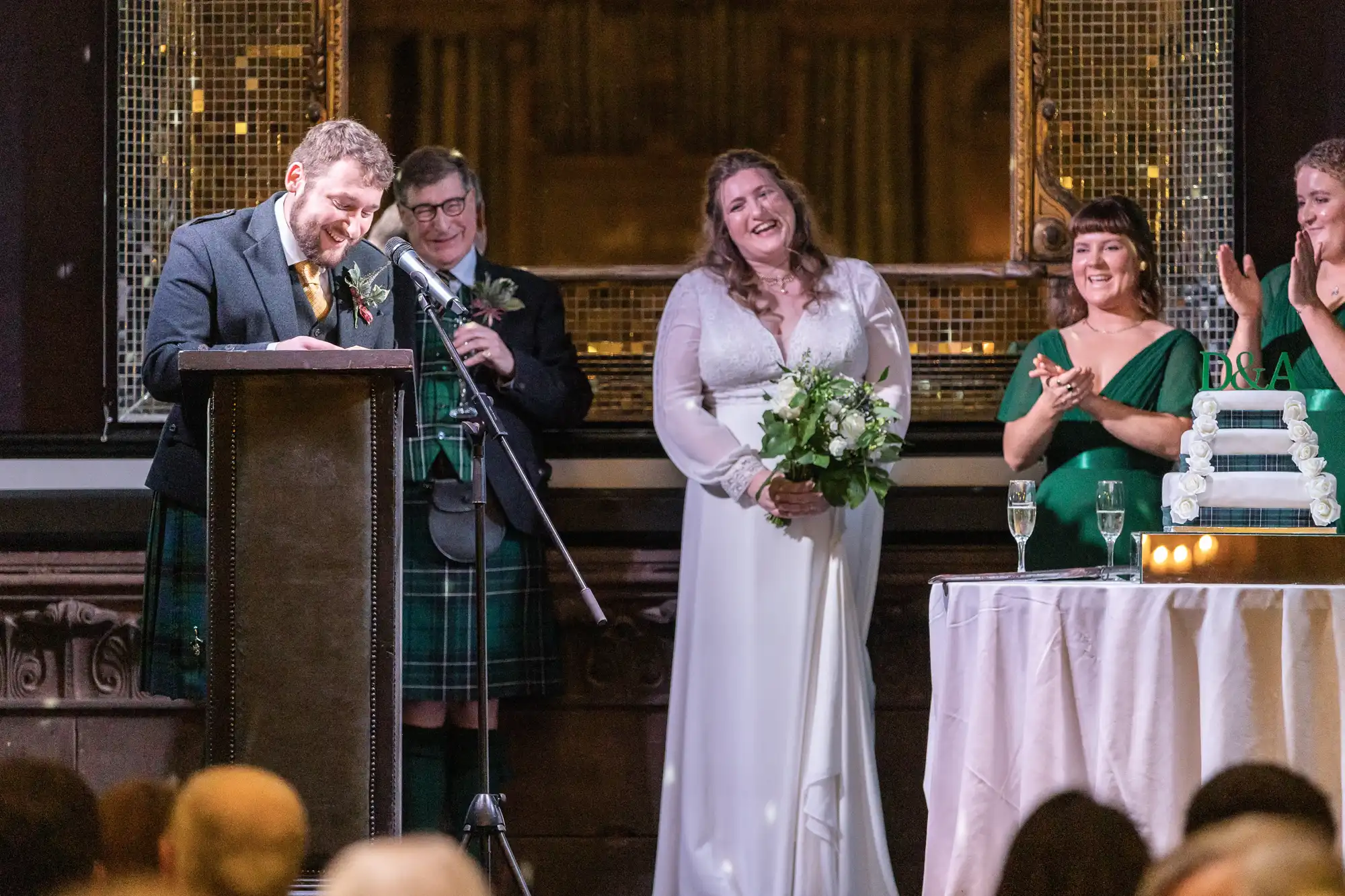 A groom speaks at a podium while the bride, holding a bouquet, smiles. Two bridesmaids in green dresses and a man in a kilt look on, standing near a table with a tiered cake and lit candles.