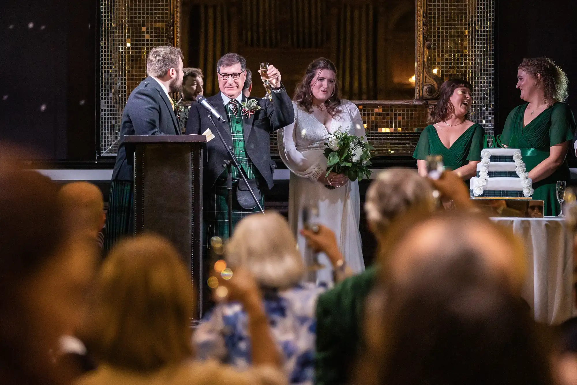 A man stands at a podium raising a glass for a toast, next to newlyweds. Guests in the foreground raise their glasses. A bride in a white dress and bridesmaids in green dresses stand near a tiered cake.