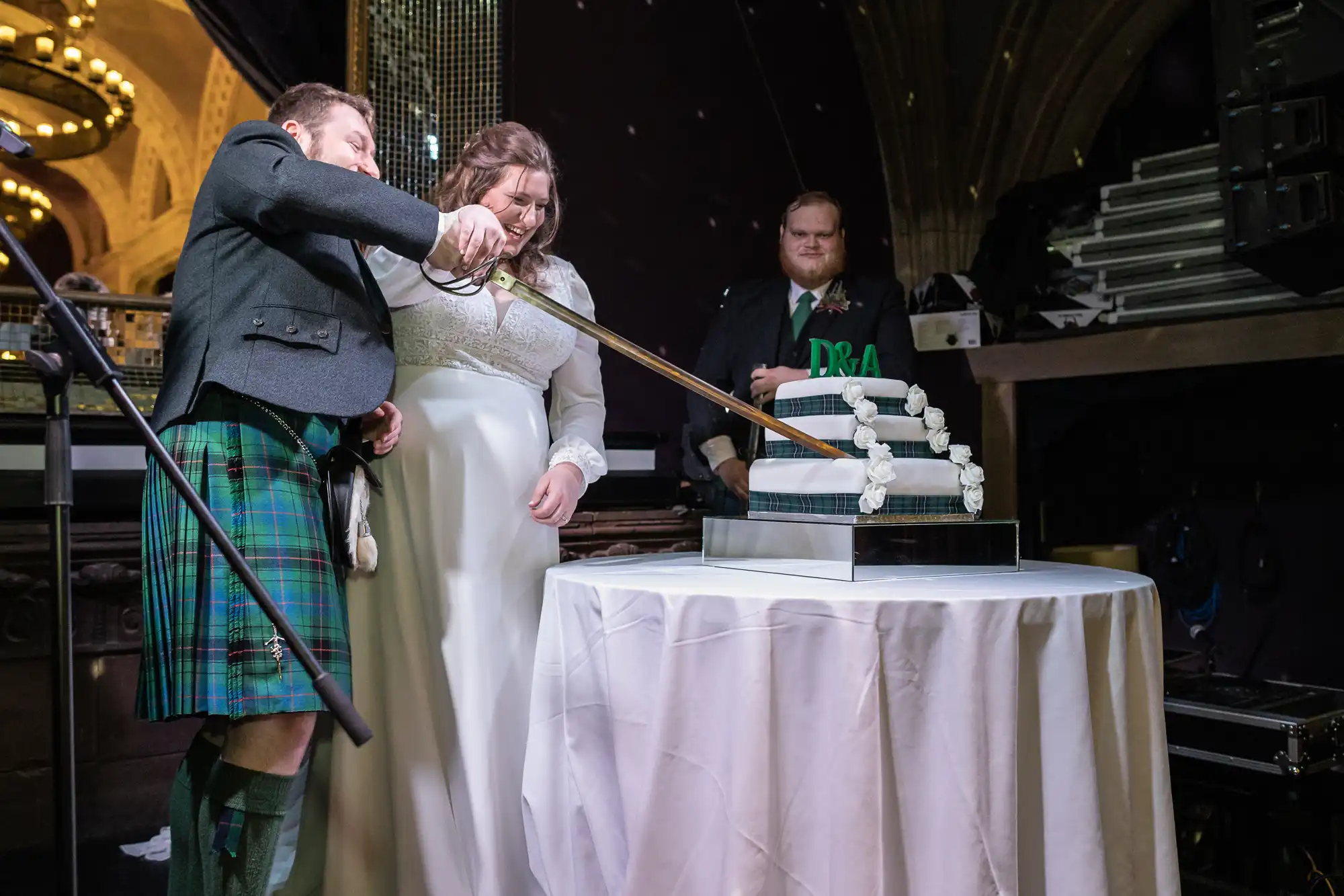 A man in a kilt and a woman in a white dress are cutting a multi-tiered wedding cake with a large sword while another man looks on.