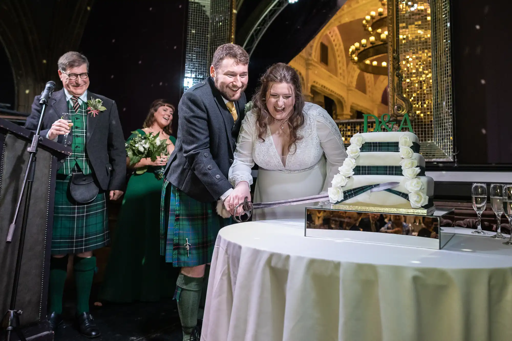 Ghillie Dhu wedding reception newlywed couple in formal attire use a sword to cut a multi-tiered wedding cake, with an audience of three people watching.