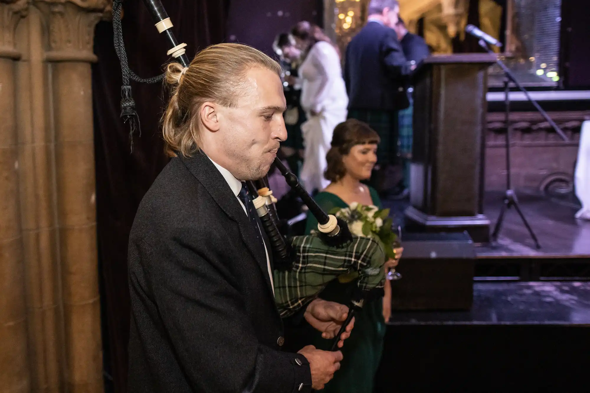 Ghillie Dhu wedding reception - A person wearing a dark jacket plays the bagpipes at an indoor event, with attendees and a podium in the background.