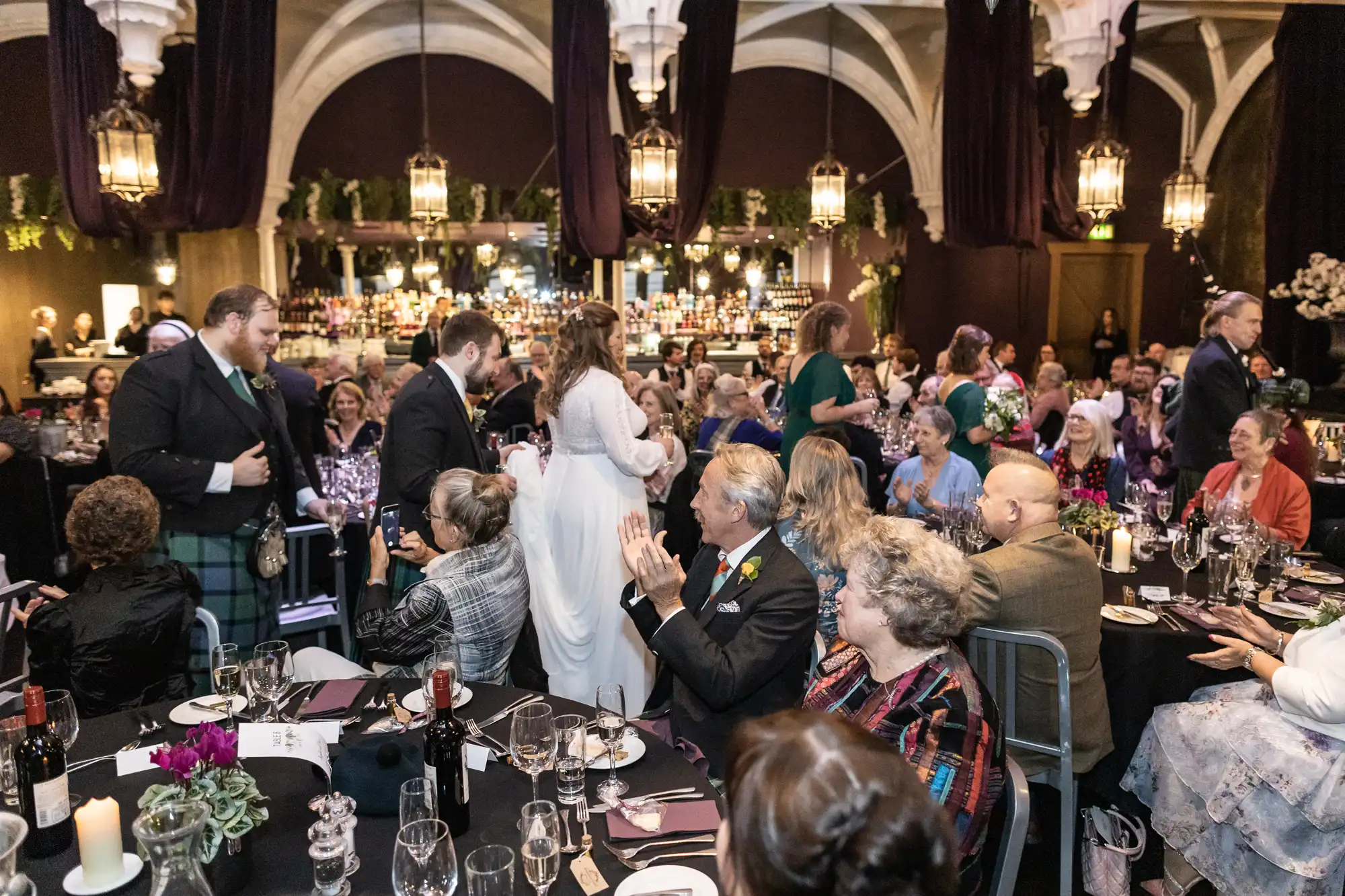 A group of people dressed in formal attire are gathered in a banquet hall. Some are seated at tables with food and drinks, while a couple in wedding attire walks past. The setting is elegant and festive.