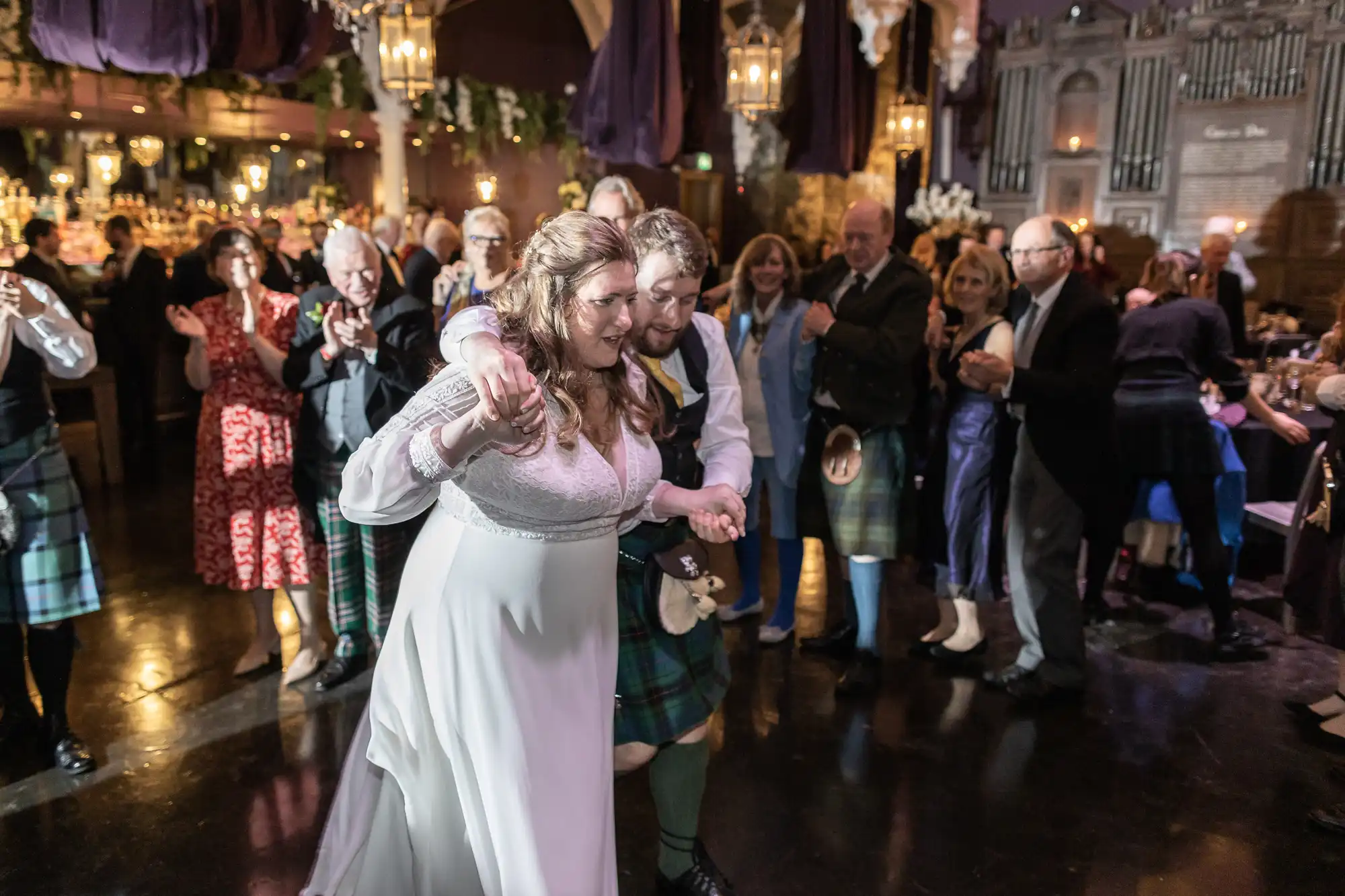 A bride and groom in traditional Scottish attire dance at their wedding, surrounded by guests in a decorated hall.