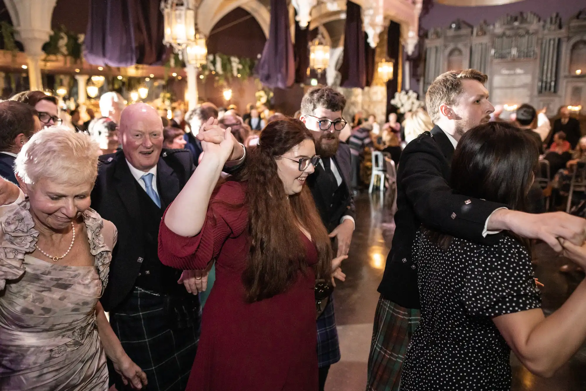 A group of people in formal attire participate in a dance at a brightly lit indoor event with ornate architecture in the background.