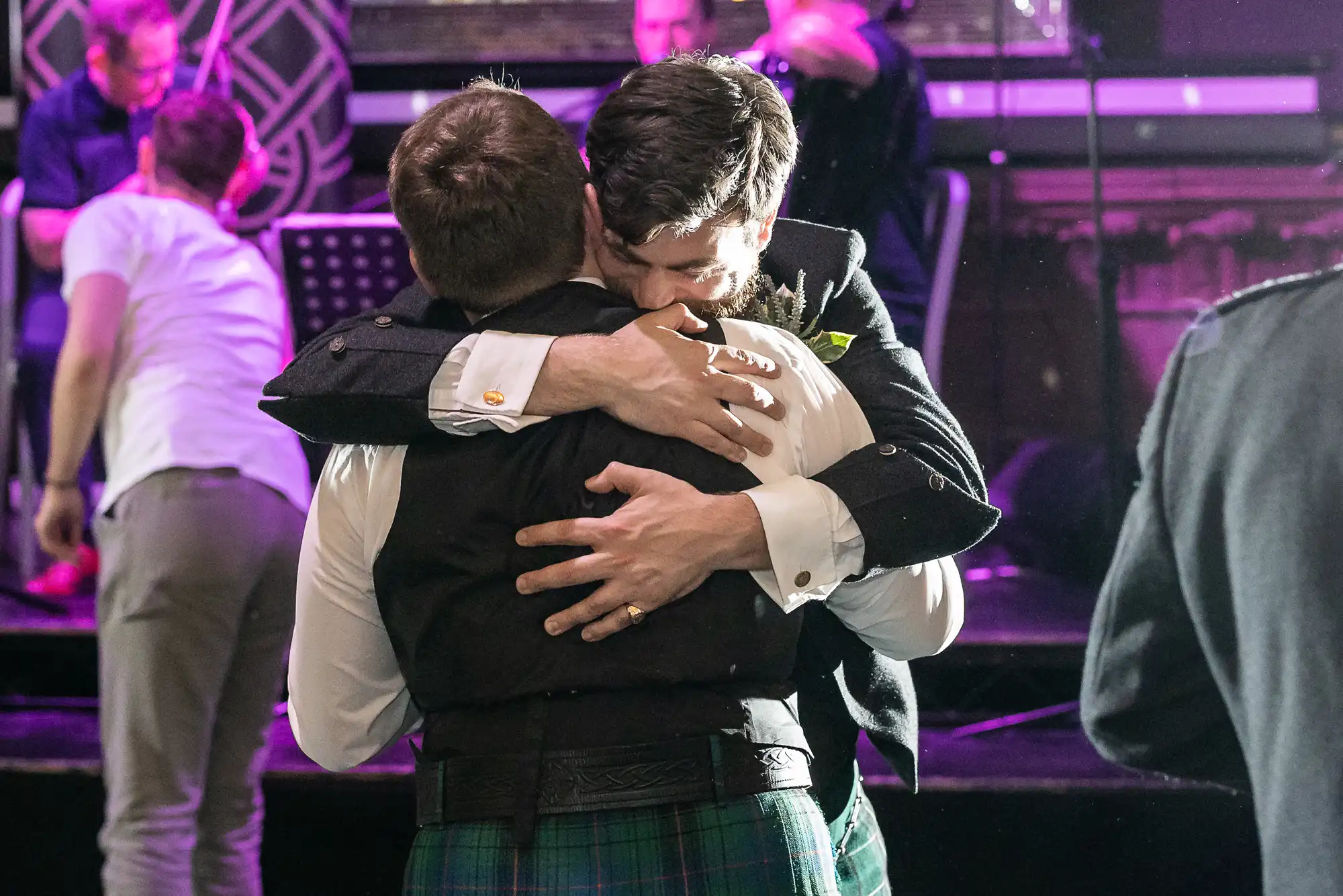 Two people wearing formal attire share a hug in what appears to be a celebratory setting with a few onlookers and musicians in the background.