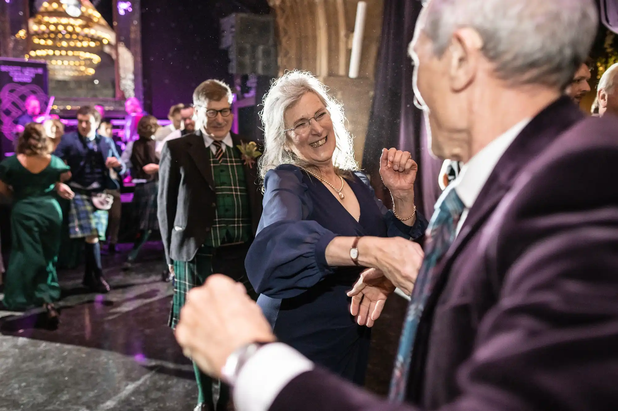 A group of people dressed in formal attire are dancing and socializing at an indoor event with colorful lights in the background.