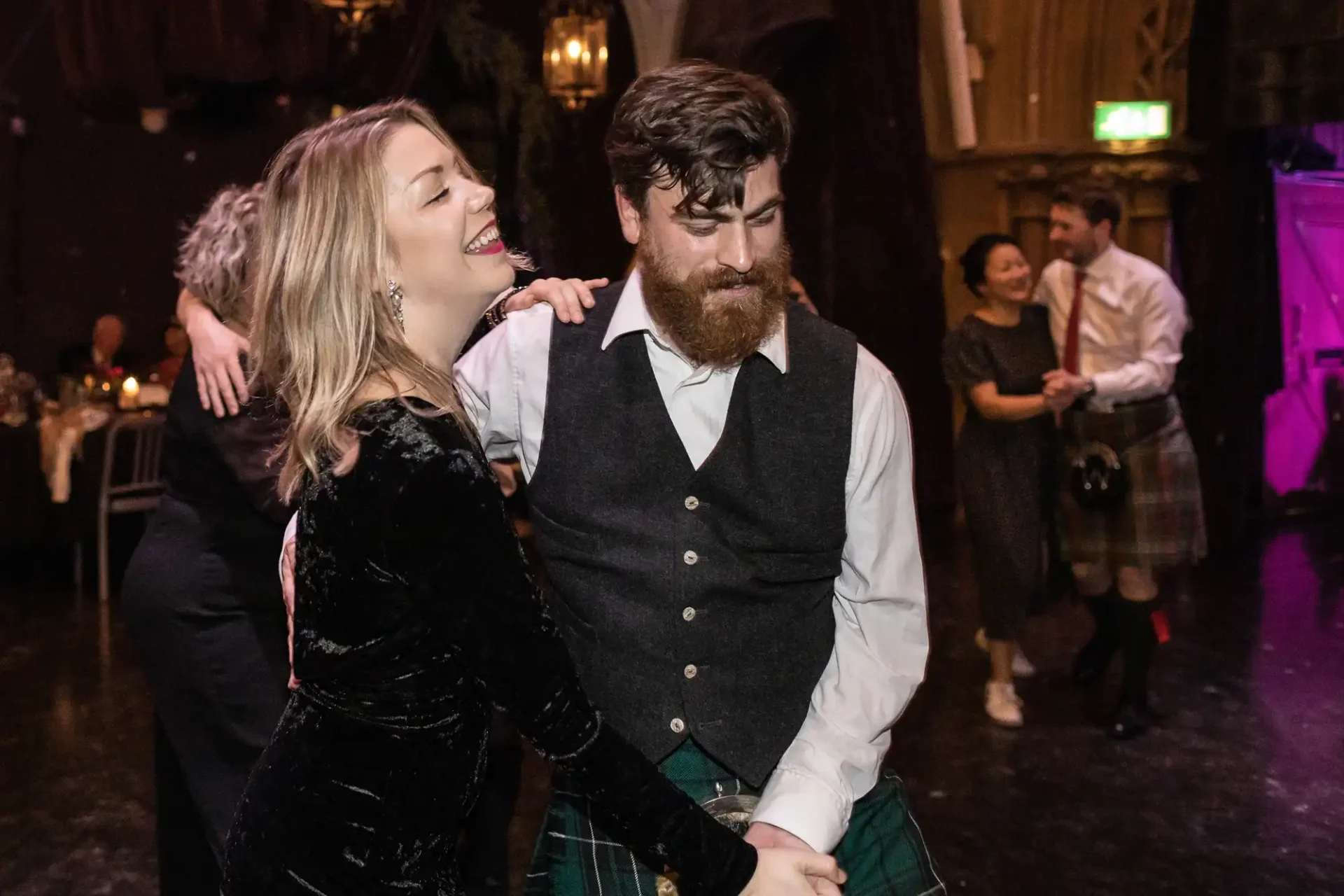 Ghillie Dhu wedding reception: A woman and man, both smiling and holding hands, dance together in a dimly lit room. Other people are dancing in the background.