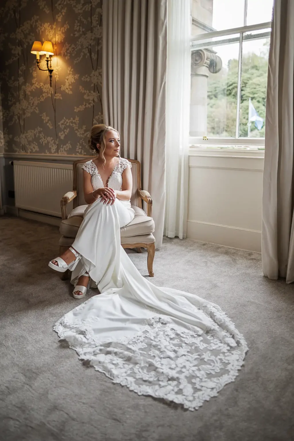A bride in a white wedding dress with a lace train sits on a chair by a window, looking outside. The room is decorated with light wallpaper and curtains.