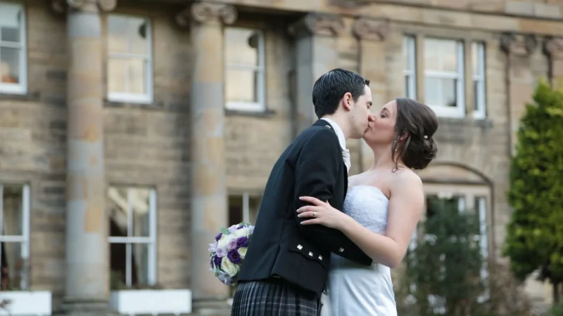 Balbirnie House wedding videographer. A bride and groom kiss outdoors in front of a stone building with columns. The groom wears a kilt and jacket; the bride wears a strapless white dress and holds a bouquet.