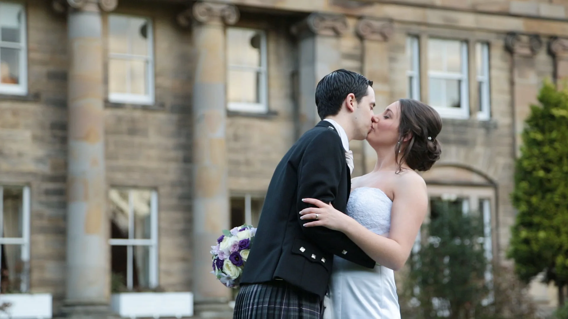 Balbirnie House wedding videographer image of a bride and groom kiss outdoors in front of a stone building with columns. The groom wears a kilt and jacket; the bride wears a strapless white dress and holds a bouquet.