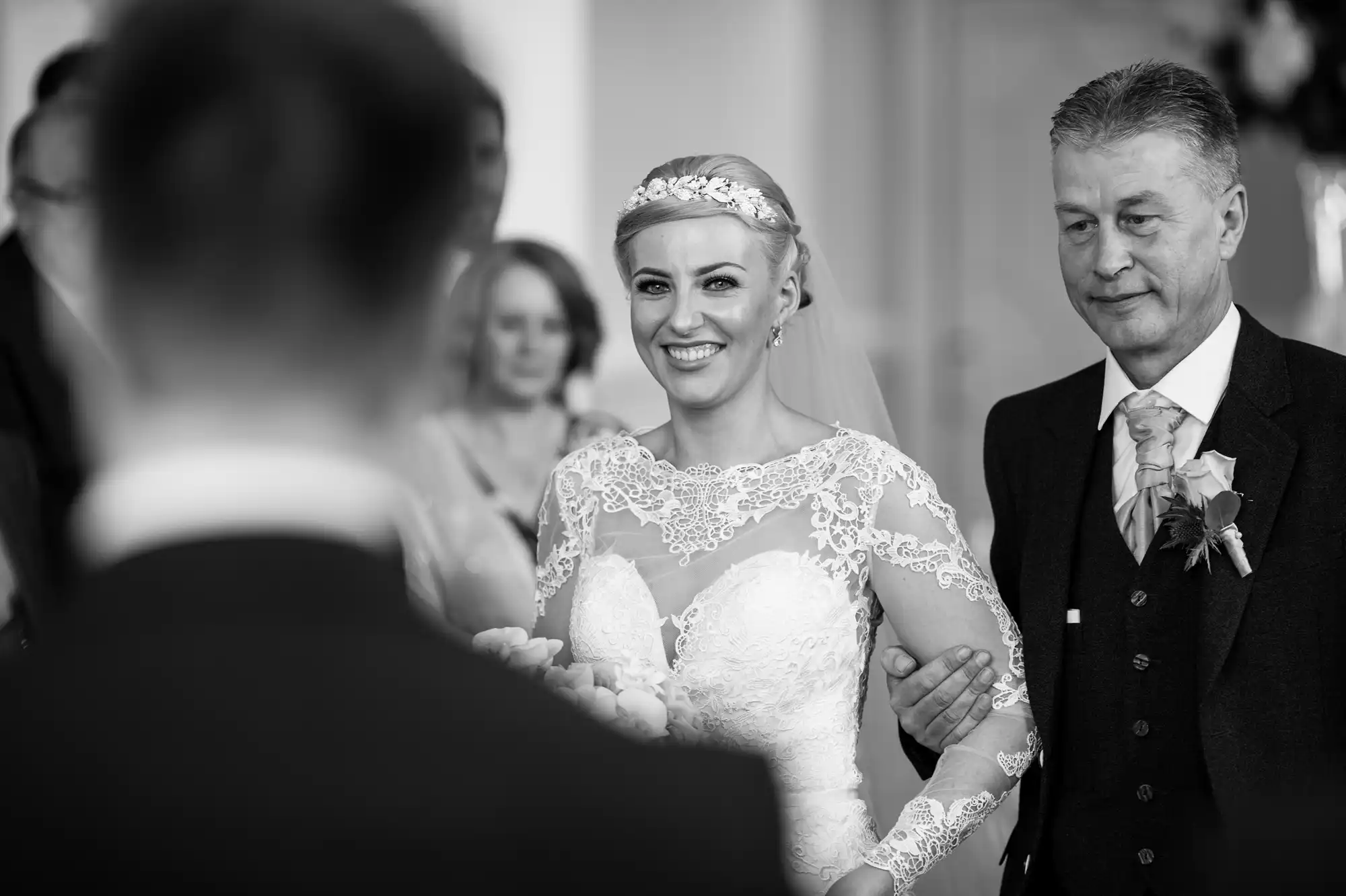 A bride in a lace wedding dress walks down the aisle with her father, smiling at her groom in the foreground. Black and white photo.