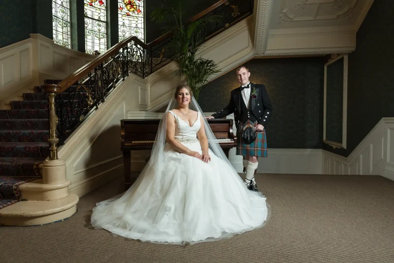 A bride in a white wedding gown sits near a piano, while the groom in a traditional Scottish kilt stands beside her on the stairs. The setting is an elegant room with stained glass windows and ornate decor.