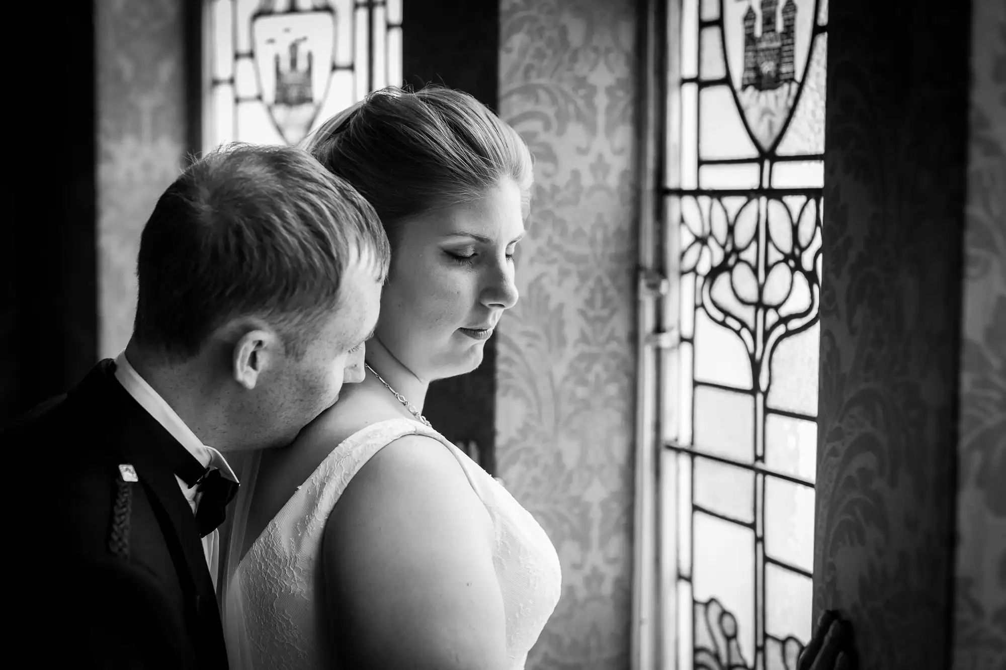 A bride and groom in wedding attire stand closely together near a window with stained glass, the groom gently resting his head on the bride's shoulder.