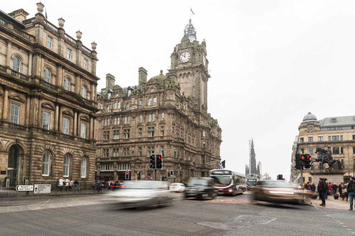 Balmoral Hotel viewed from Waterloo Place: a busy city street with vehicles in motion and pedestrians near historic stone buildings and a clock tower under a cloudy sky. Traffic lights with red signals are visible.