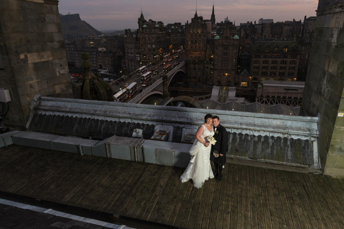 newlyweds on the roof of the Balmoral Hotel at dusk with Edinburgh Old Town in the background