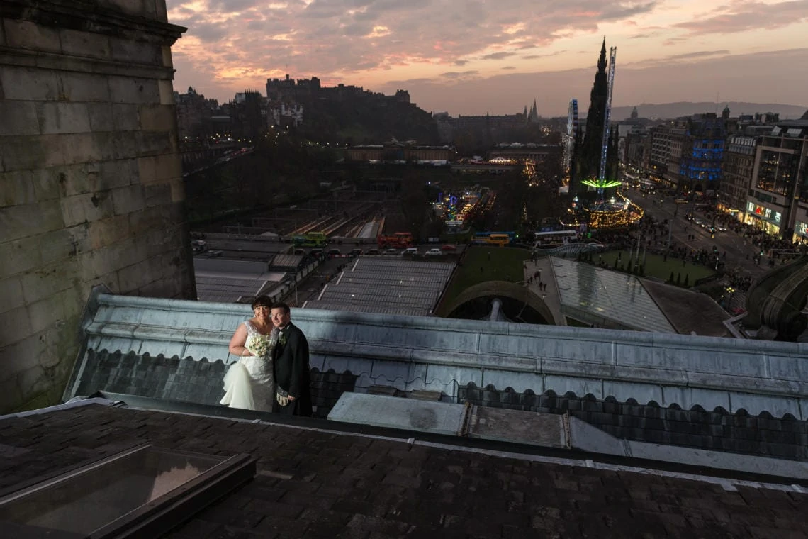 newlyweds on the roof of the Balmoral Hotel at dusk with Edinburgh Castle and Scott Monument in the background
