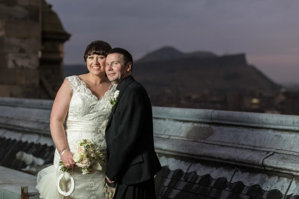 newlyweds on the roof of the Balmoral Hotel at dusk with Arthur's Seat in the background