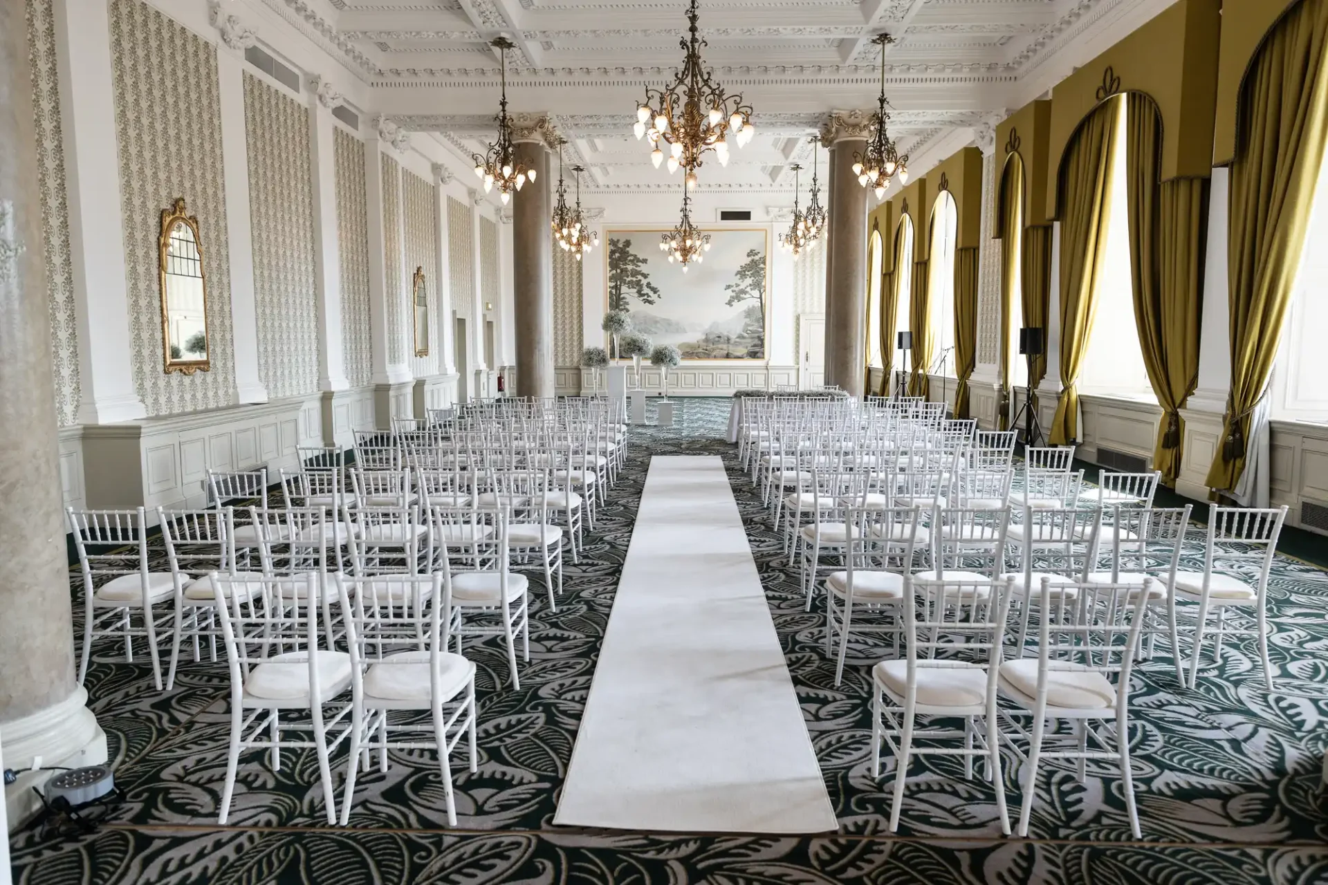 Elegant wedding venue with rows of white chairs arranged on either side of a long white aisle runner, leading to a large window, in a room with ornate chandeliers and drapery.