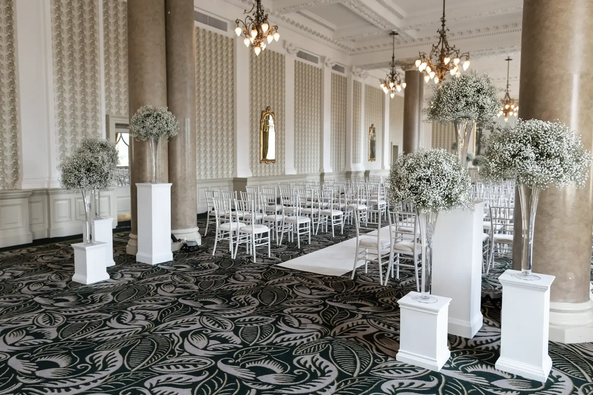 Elegant indoor wedding venue setup with rows of white chairs, floral arrangements on pedestals, and chandeliers in a room with patterned carpet and columned walls.