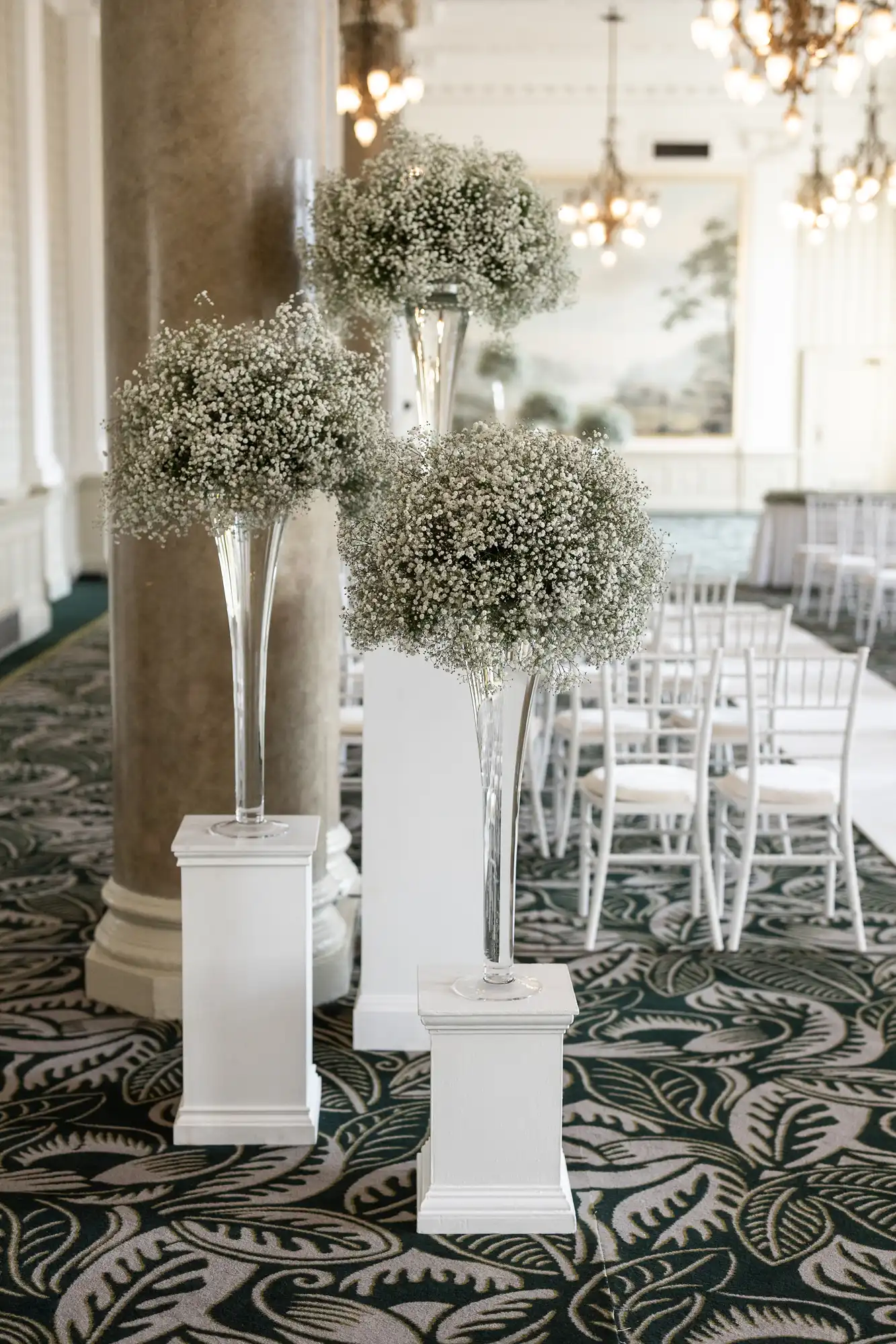 Three tall floral arrangements on white pedestals in an elegant room with ornate chandeliers and patterned carpet.