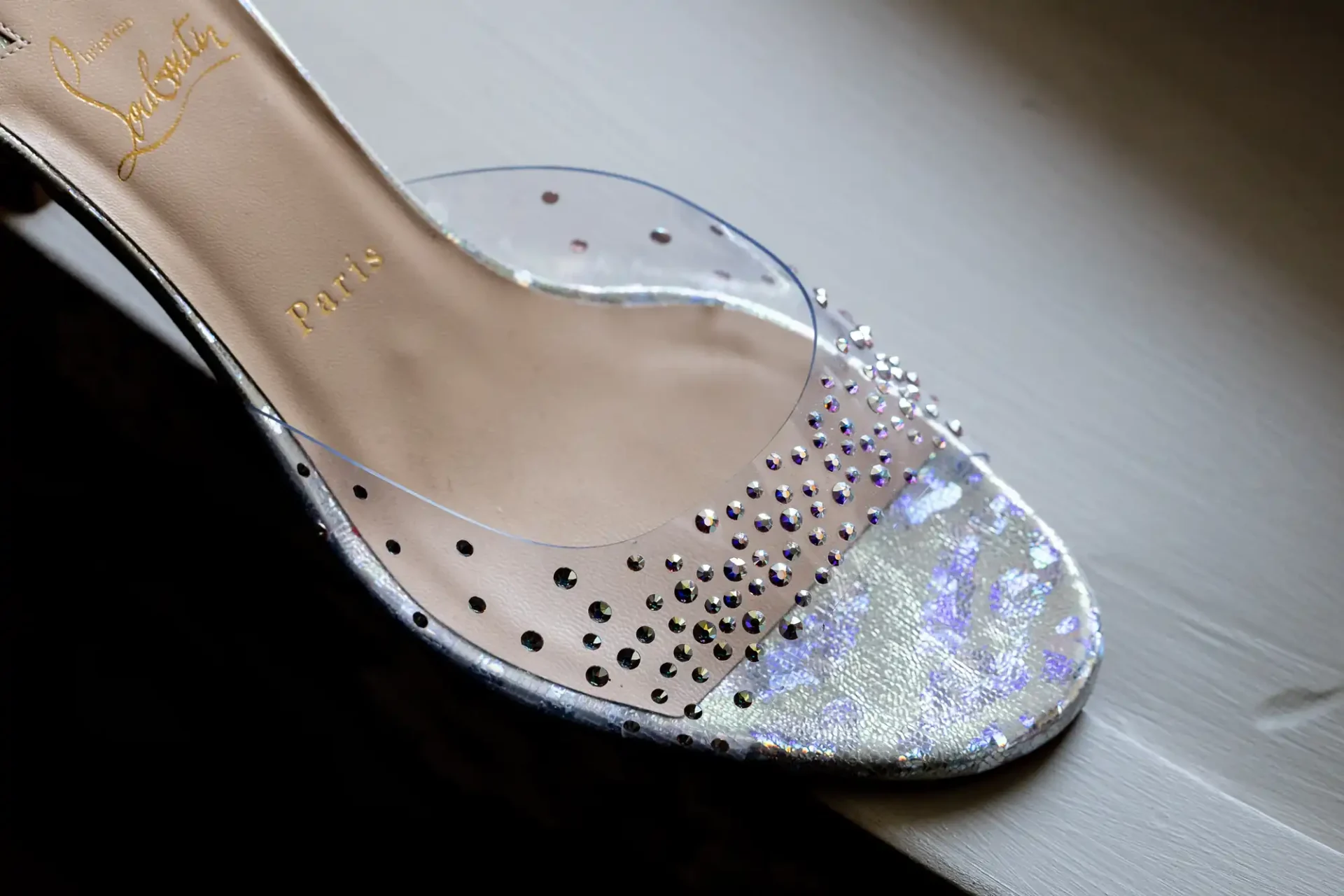 A close-up of a christian louboutin transparent slipper with rhinestone embellishments, displayed on a wooden surface.