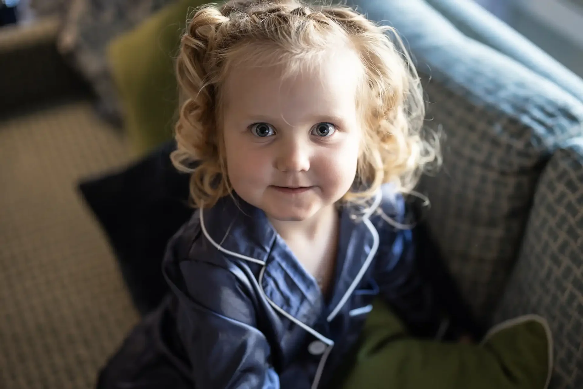 A toddler with curly blonde hair smiling while wearing a blue jacket, seated indoors on a cushioned surface.