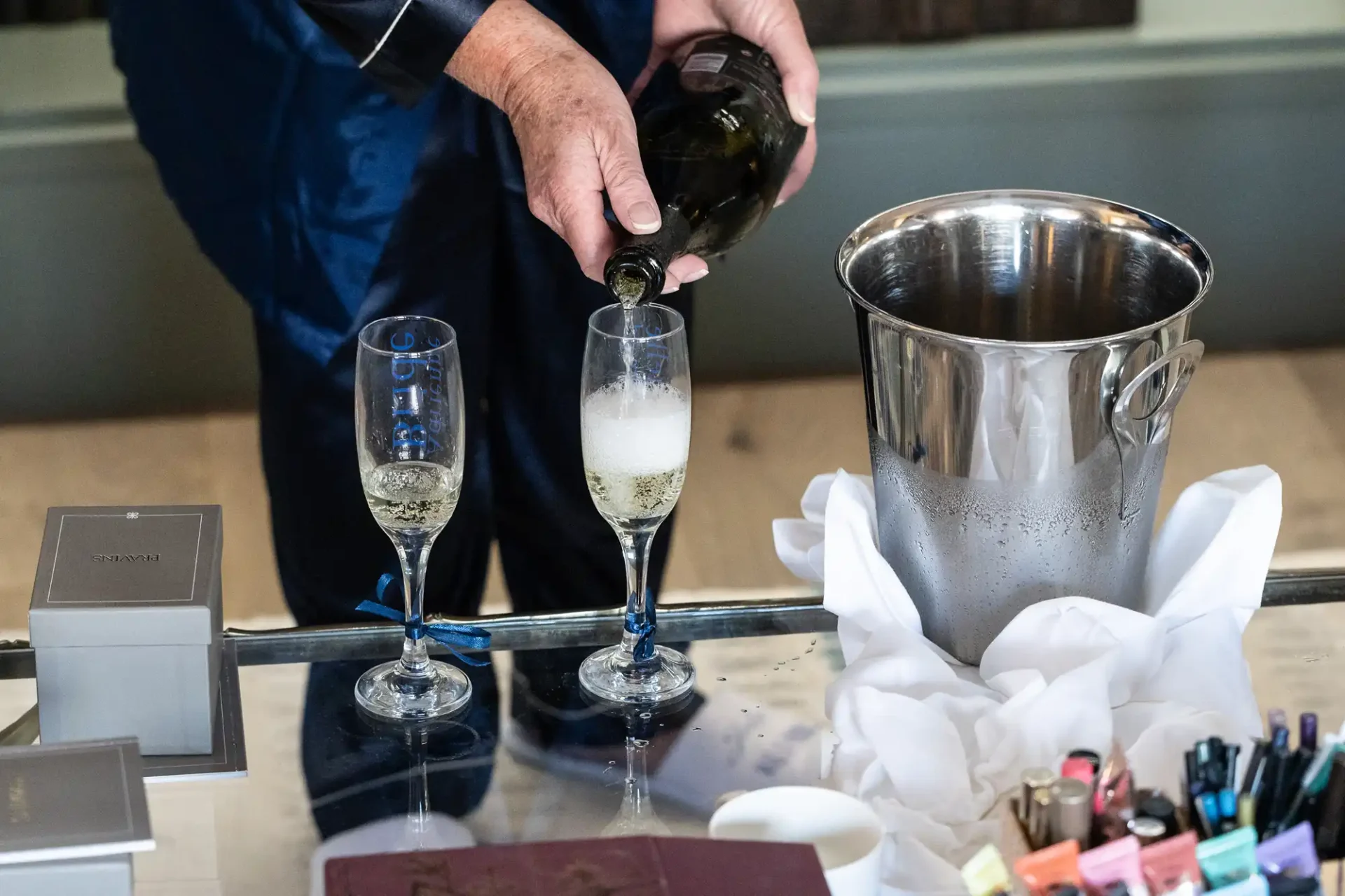 A person pours champagne into two flutes on a table with a silver ice bucket and various colored pens nearby.