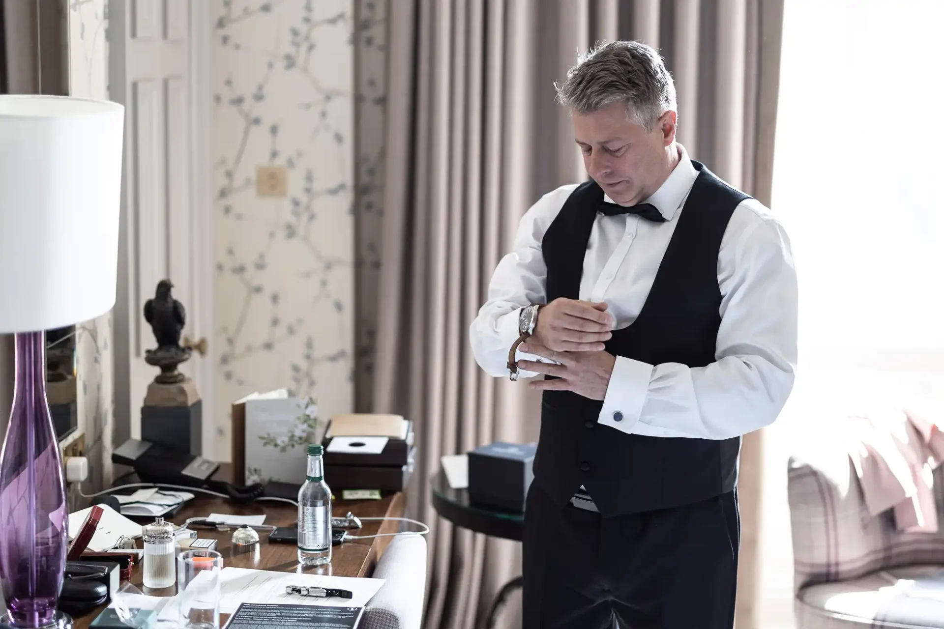 A man in a tuxedo adjusts his cufflinks in an elegantly furnished room with a desk and decor items.