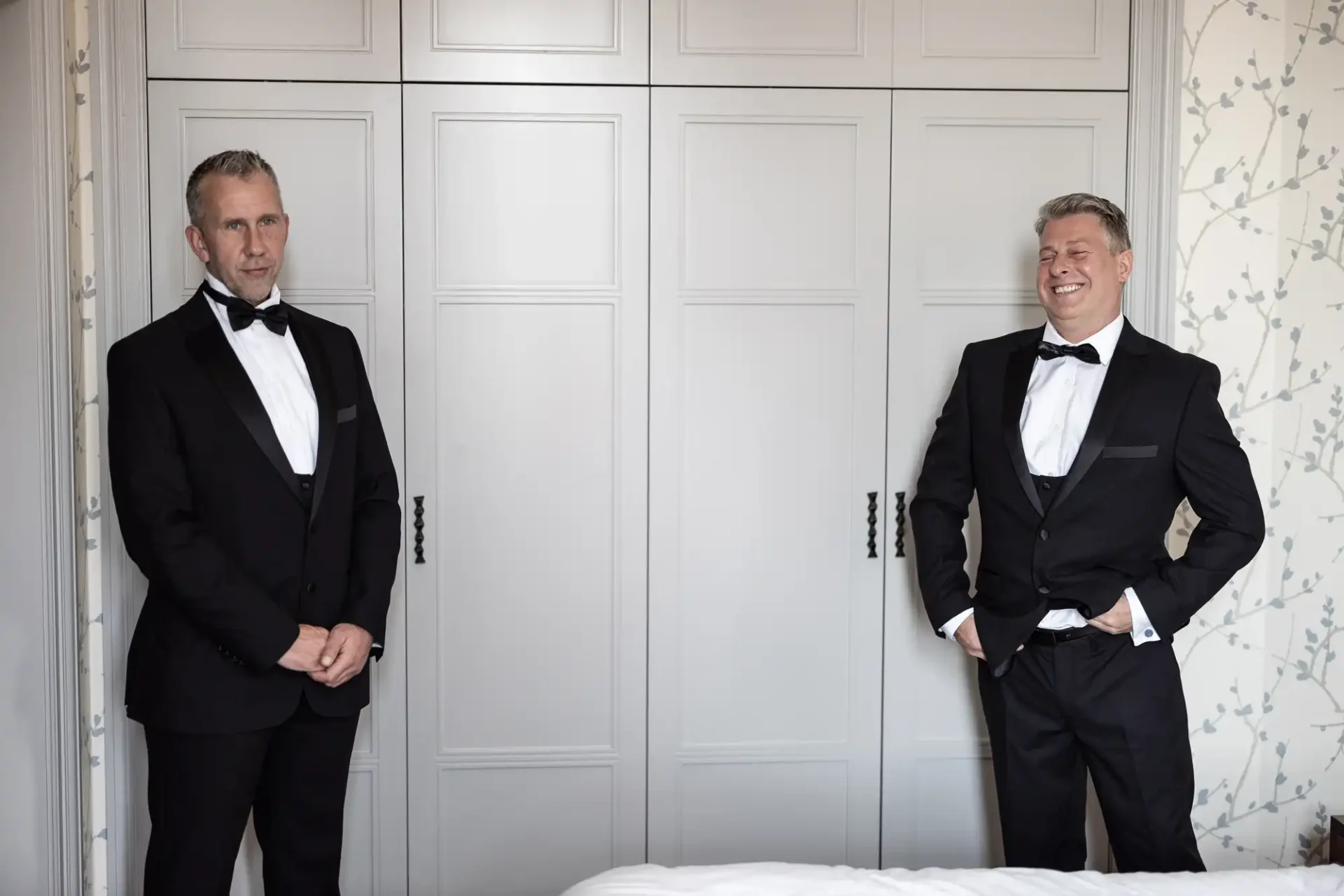 Two men in tuxedos smiling and standing apart against a white paneled wall in an elegantly decorated room.
