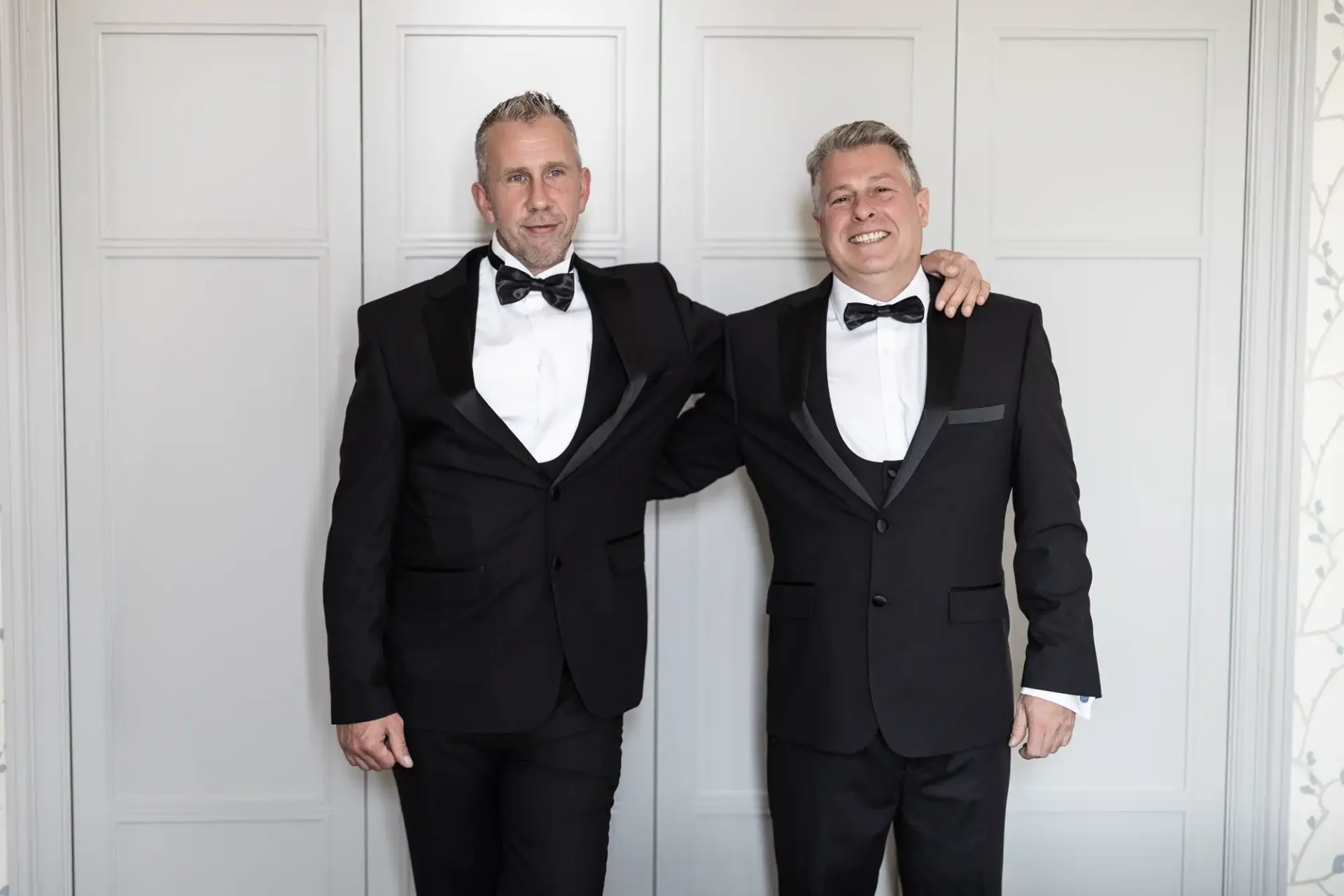 Two smiling men in tuxedos standing arm in arm in a room with white paneled walls.