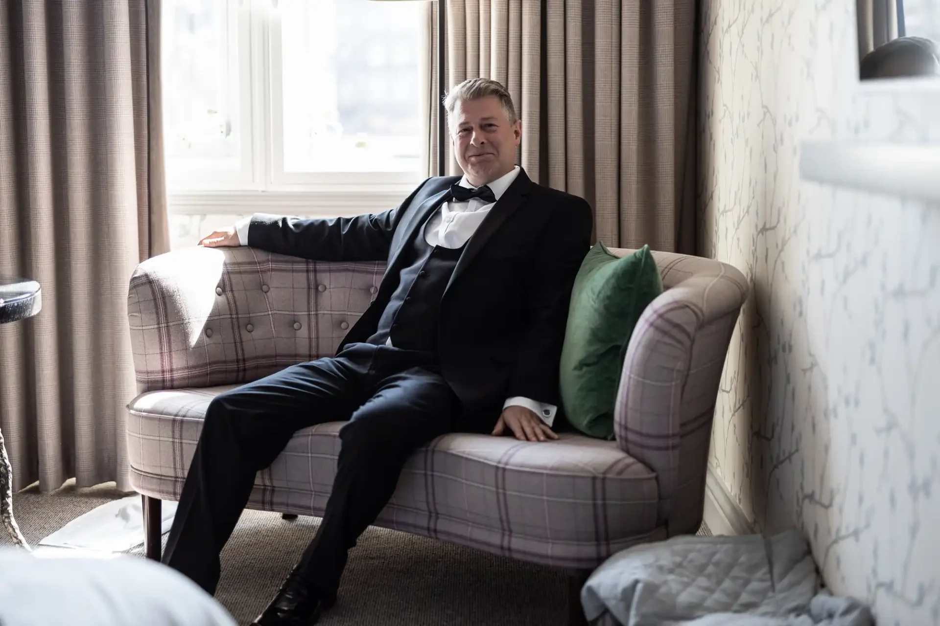 Man in a black suit sitting relaxedly on a pink couch by a window in a well-lit room with curtains.