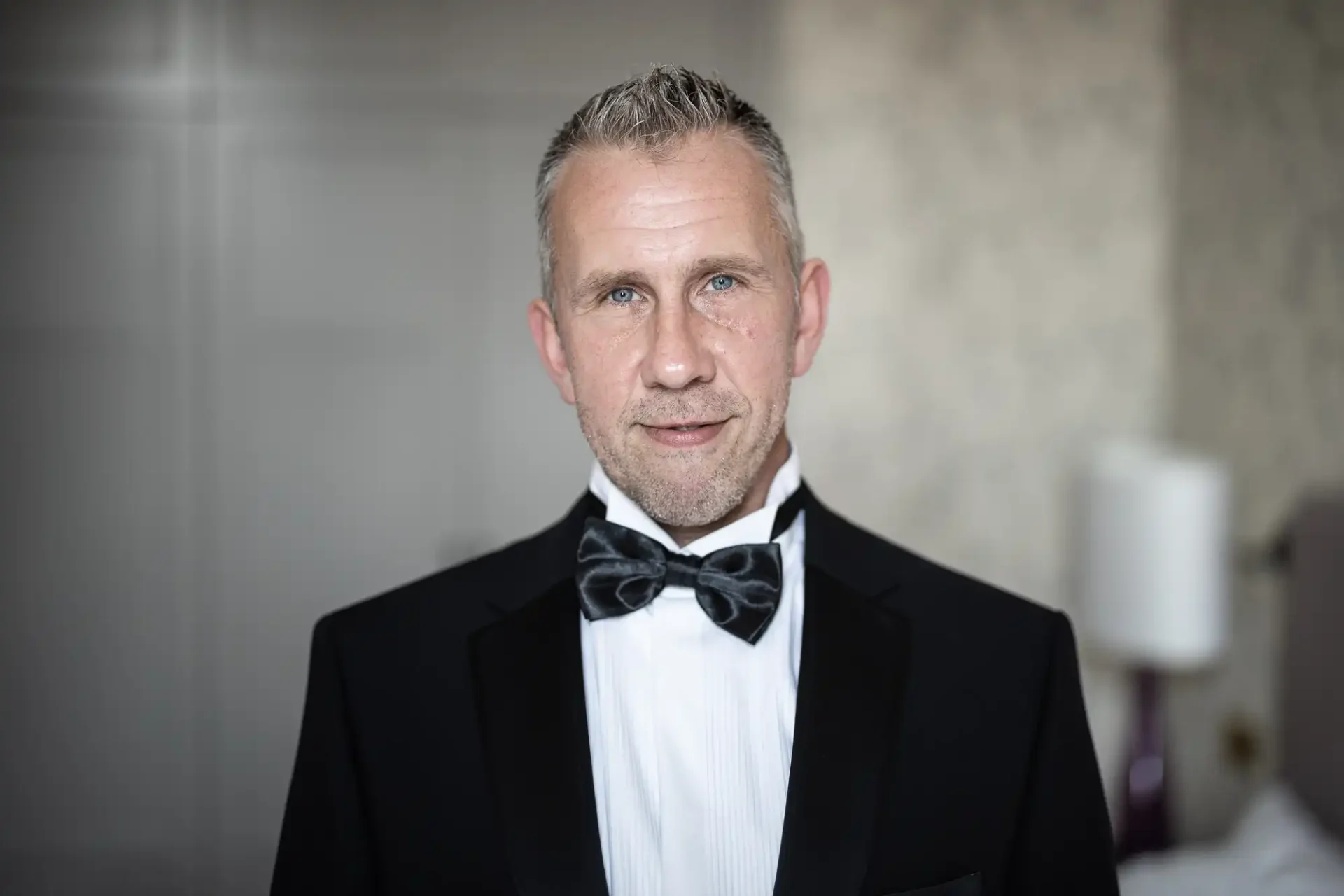A middle-aged man with gray hair, wearing a black tuxedo and bow tie, standing in a well-lit room.