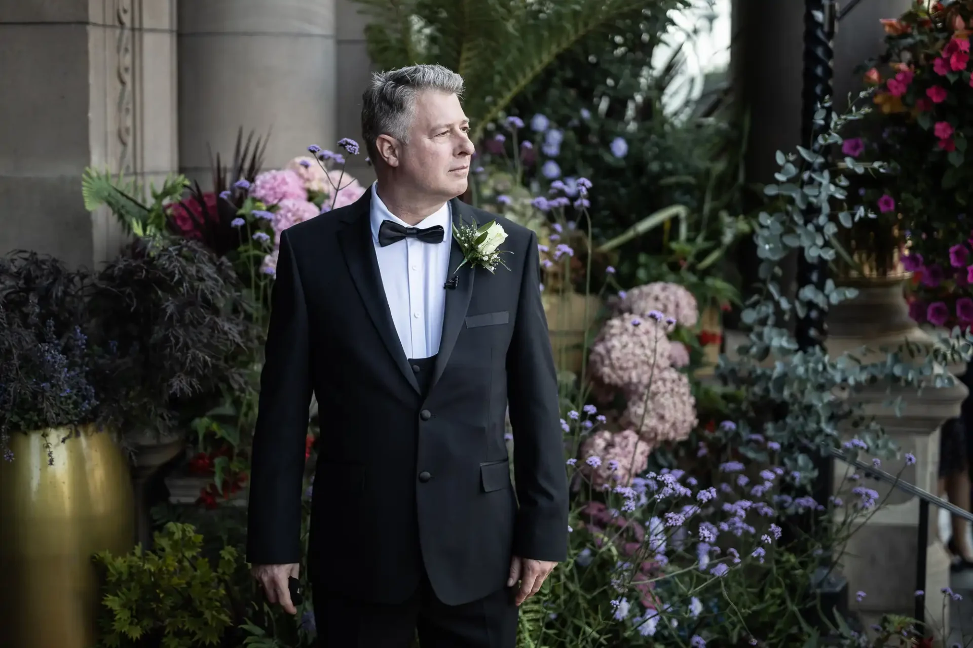Man in a tuxedo with a boutonniere standing by a floral display at a formal event.