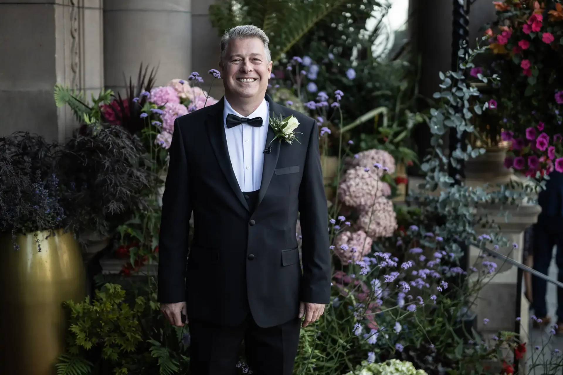 A smiling man in a black tuxedo and bow tie stands in front of a floral arrangement.