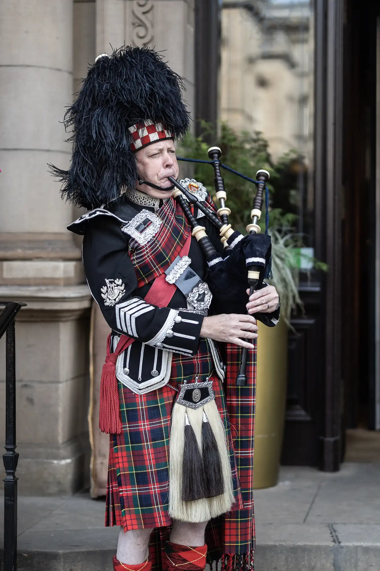 A bagpiper in traditional scottish attire, featuring a tartan kilt and a feathered bonnet, plays the bagpipes on a city street.