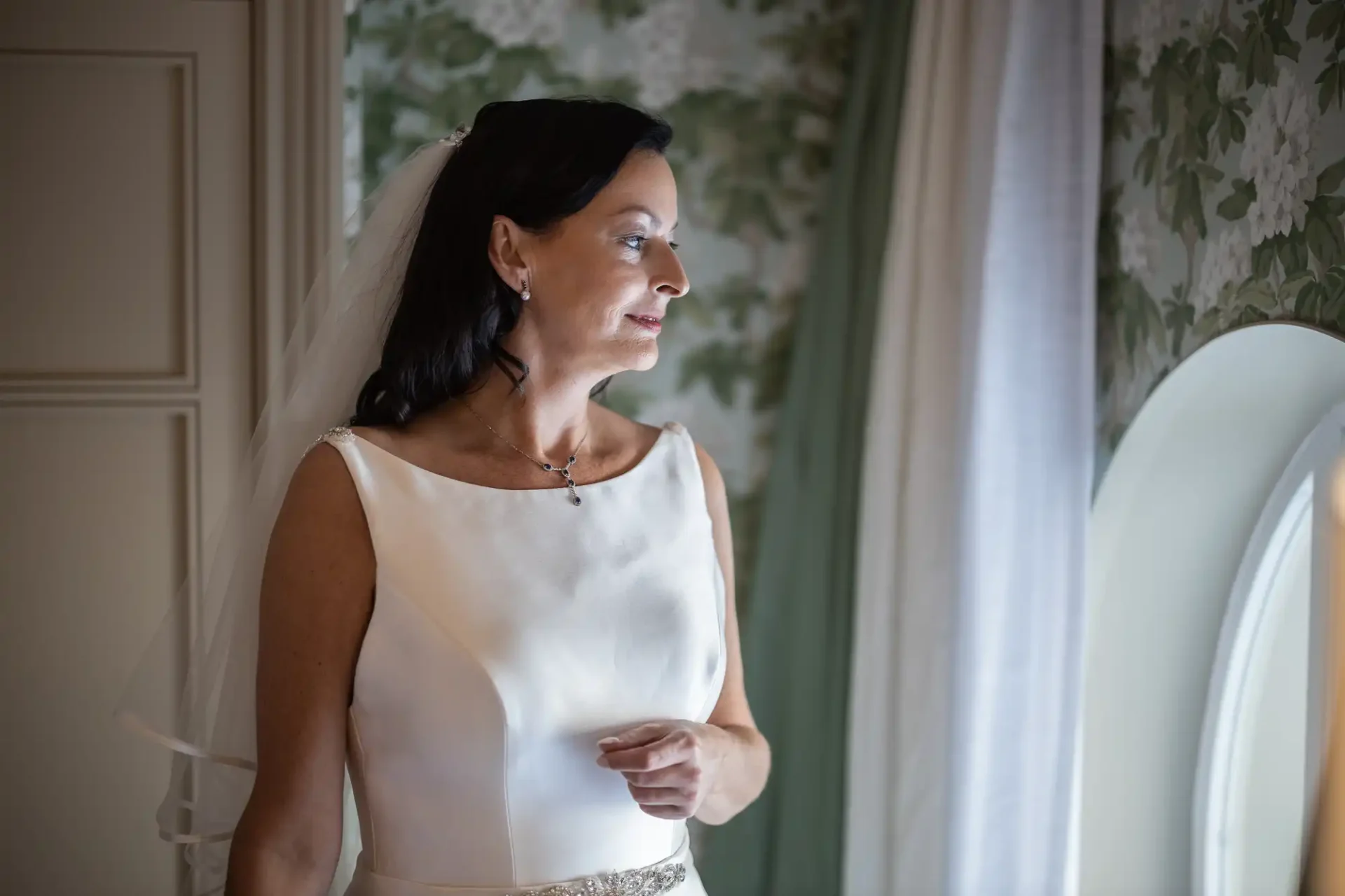 A mature bride in a white dress and veil looks at a mirror, her expression thoughtful and serene.