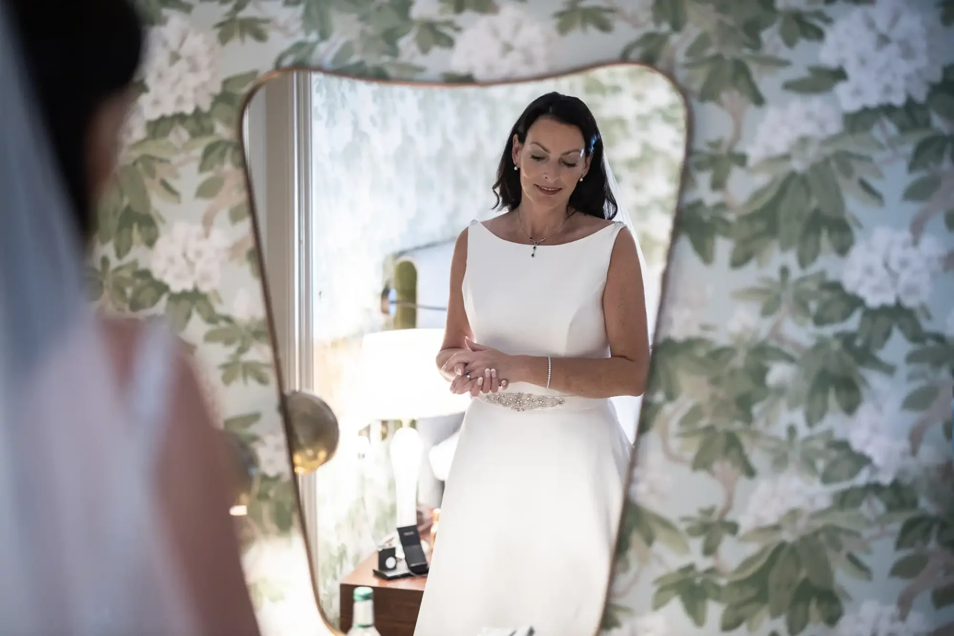 A woman in a white dress adjusts her bracelet while looking at her reflection in a mirror framed by floral wallpaper.