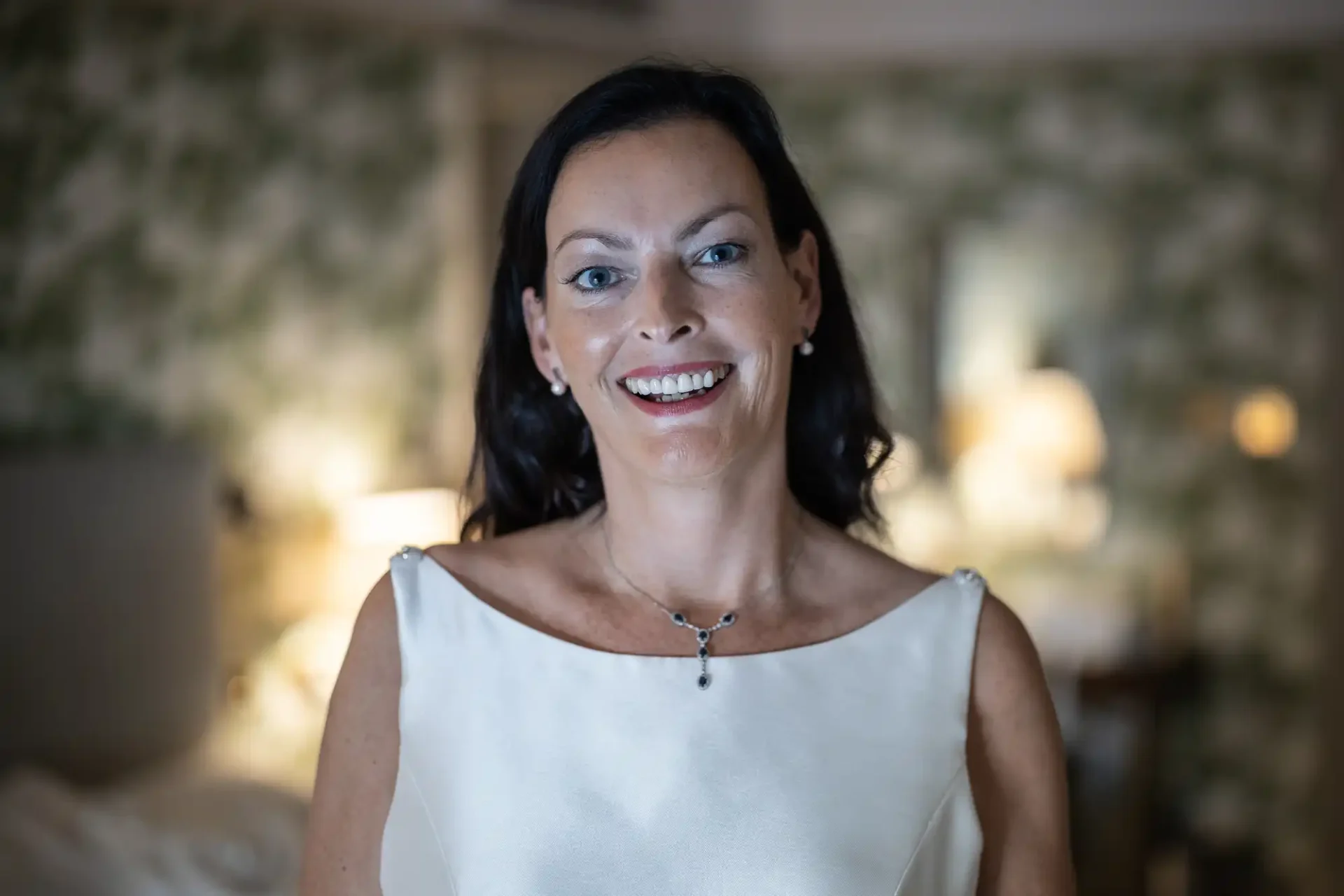A smiling woman with dark hair, wearing a sleeveless white dress and a pendant necklace, stands in a softly lit room with floral wallpaper.