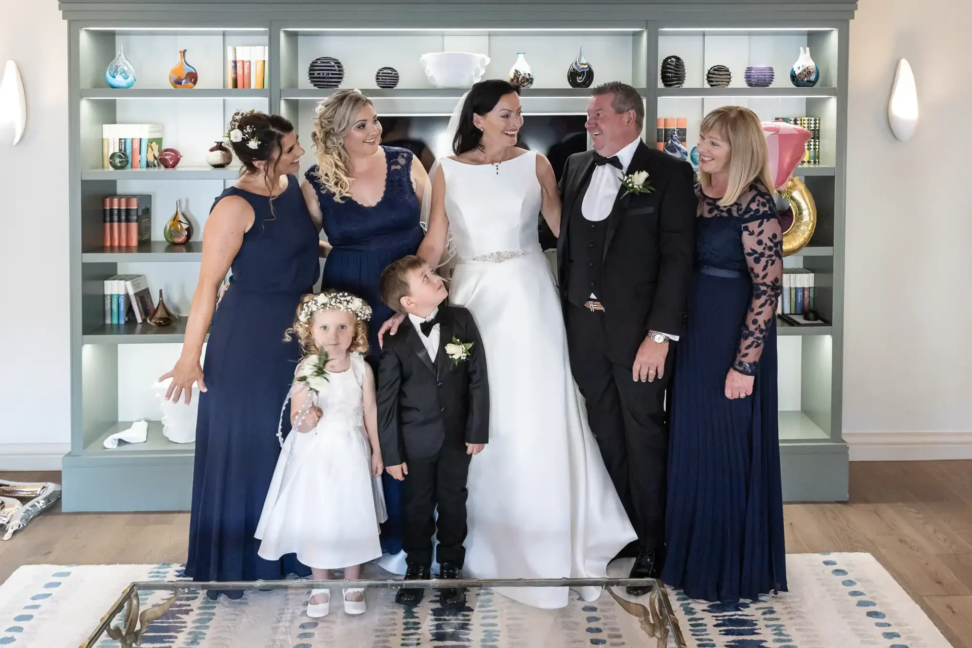 A joyful wedding group indoors, featuring two children in the center with adults in formal attire laughing around them.