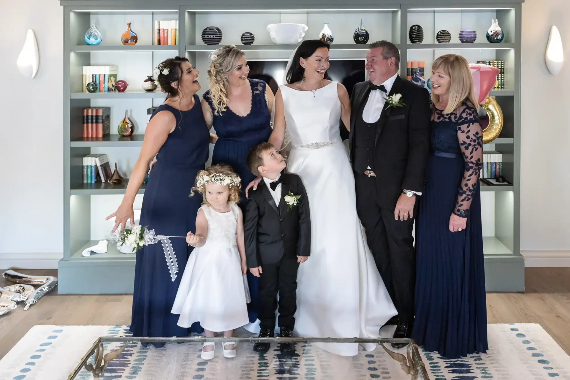 A happy wedding party inside a living room with a bride, groom, three adult women, and two children in formal attire, all smiling, in front of a shelved backdrop with decorative items.