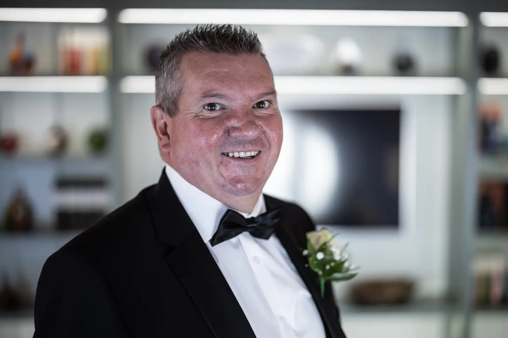 A smiling man in a tuxedo with a boutonniere, standing in a room with a blurry background featuring shelves and a tv.