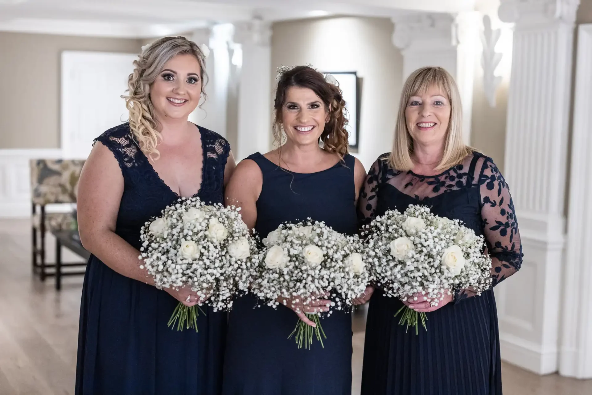 Three women in dark blue dresses holding large bouquets of white flowers, smiling in an elegant room with white columns.