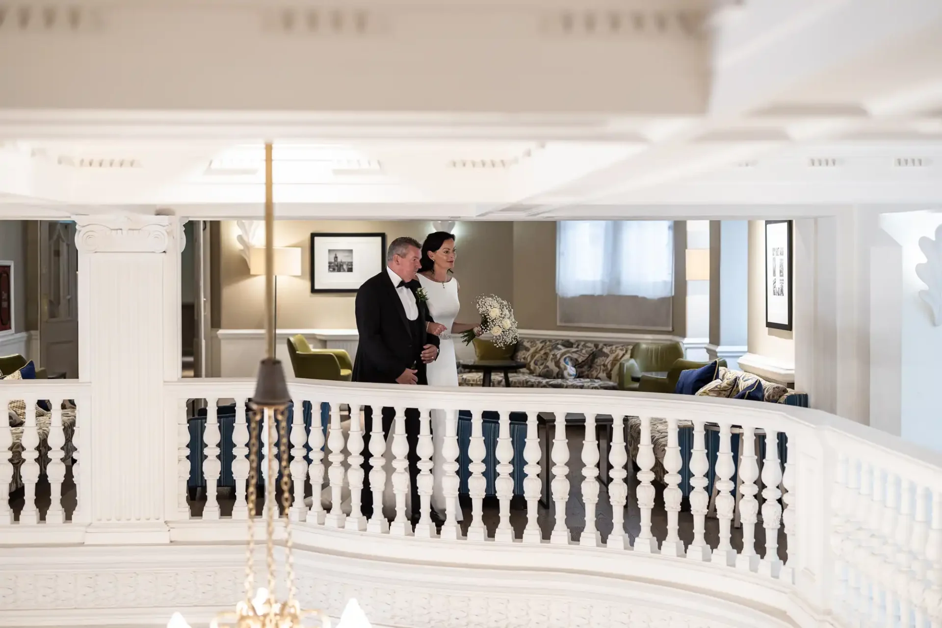 A couple dressed in formal attire stands on an elegant white balcony, overlooking a classy interior setting.