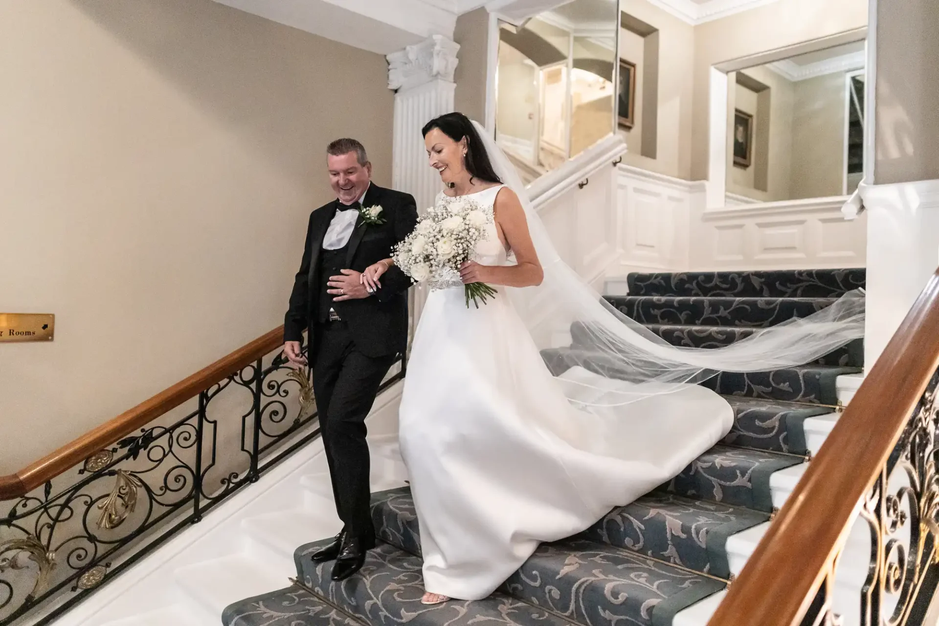 A bride in a white gown and a groom in a black suit descend a staircase, smiling joyfully, with the bride holding a bouquet.